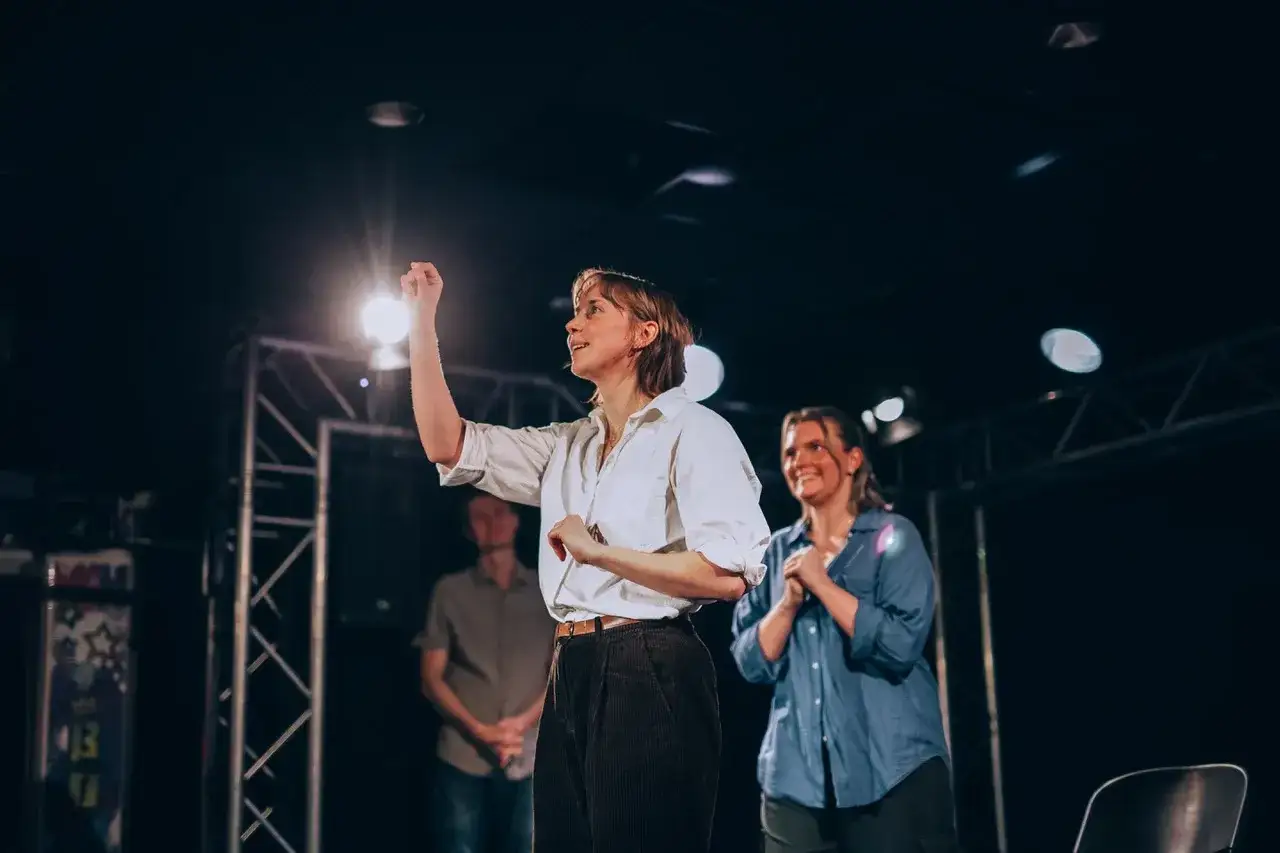 Scen från en improv-föreställning i Stockholm. En kvinna med vit skjorta gestikulerar, medan en annan kvinna ler bakom henne.