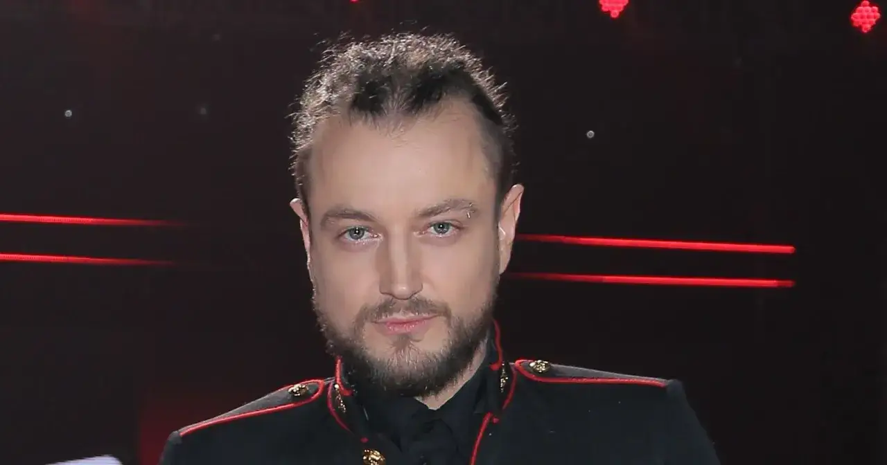 Alek Baron, znany z charakterystycznej fryzury i kolczyka w brwi, w czarnym stroju z czerwonymi zdobieniami. Jego wiek nie jest widoczny na pierwszy rzut oka.