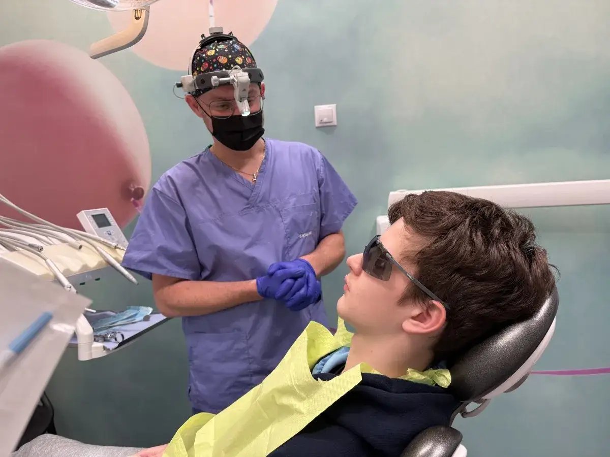 Dentysta w masce i okularach ochronnych przygotowuje się do zabiegu, być może z użyciem abrazji powietrznej, nad młodym pacjentem w okularach ochronnych.