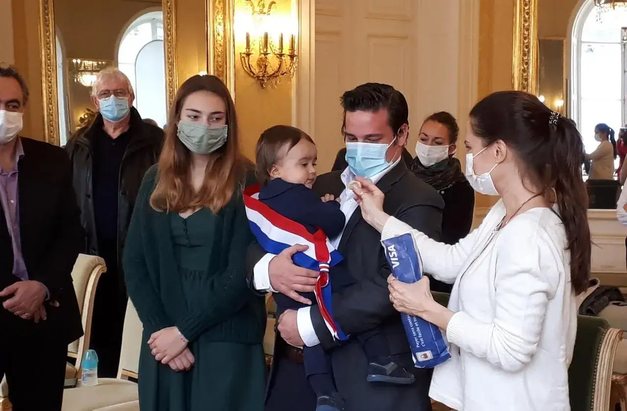 Cérémonie de baptême civil. Un bébé porte une écharpe tricolore. La démarche est solennelle.