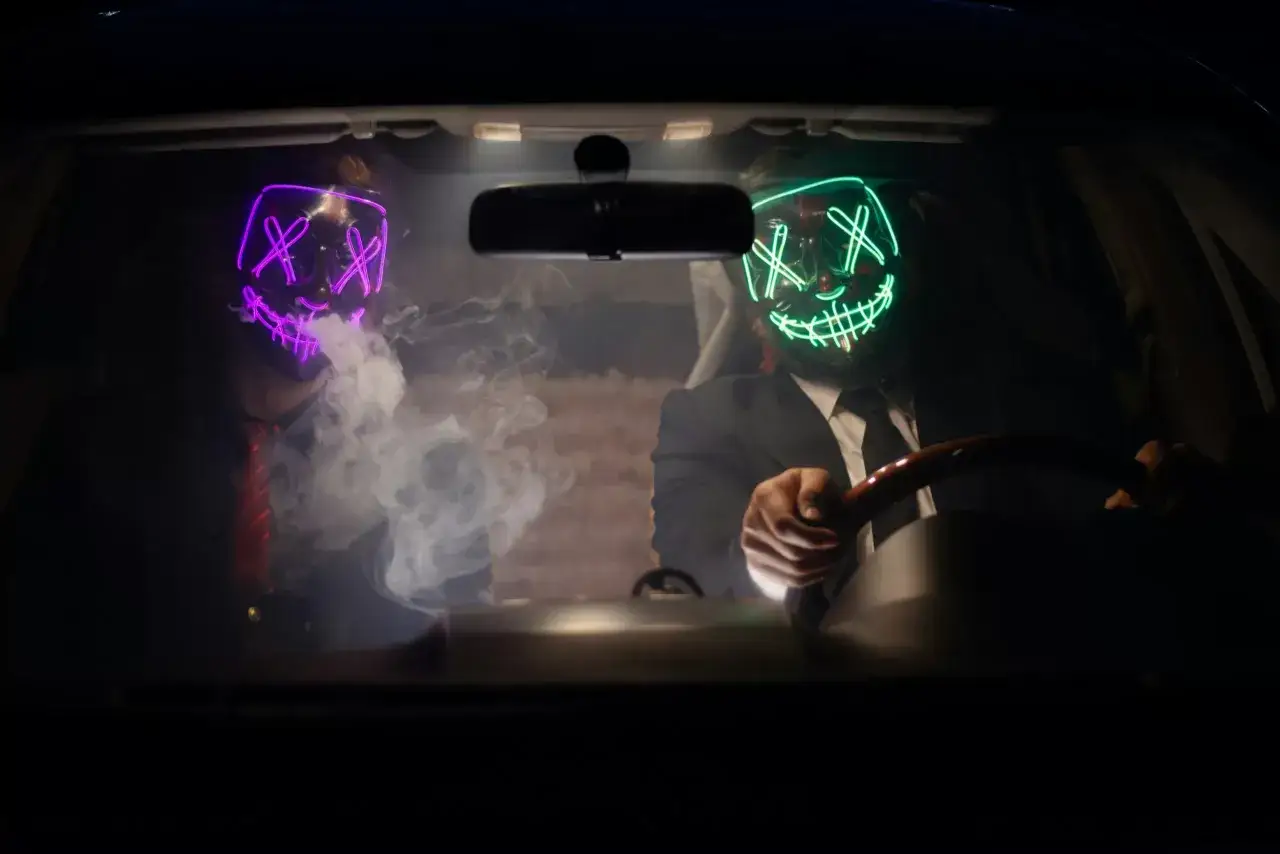 Dwóch mężczyzn w neonowych maskach, jeden z fioletową, drugi z zieloną, siedzi w samochodzie. Dym unosi się wokół nich, sugerując mroczne zamiary, być może związane z cyberprzestępczością.
