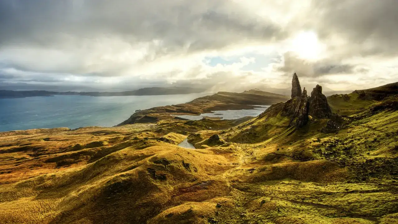 Dramatische Felsformationen ragen aus den gr&uuml;nen H&uuml;geln der Schottland Highlands. Ein See glitzert im Licht, das durch die Wolken bricht.
