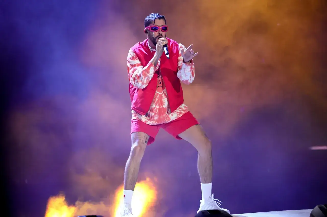 Bad Bunny w czerwonym stroju i okularach śpiewa na scenie, otoczony dymem i światłami.