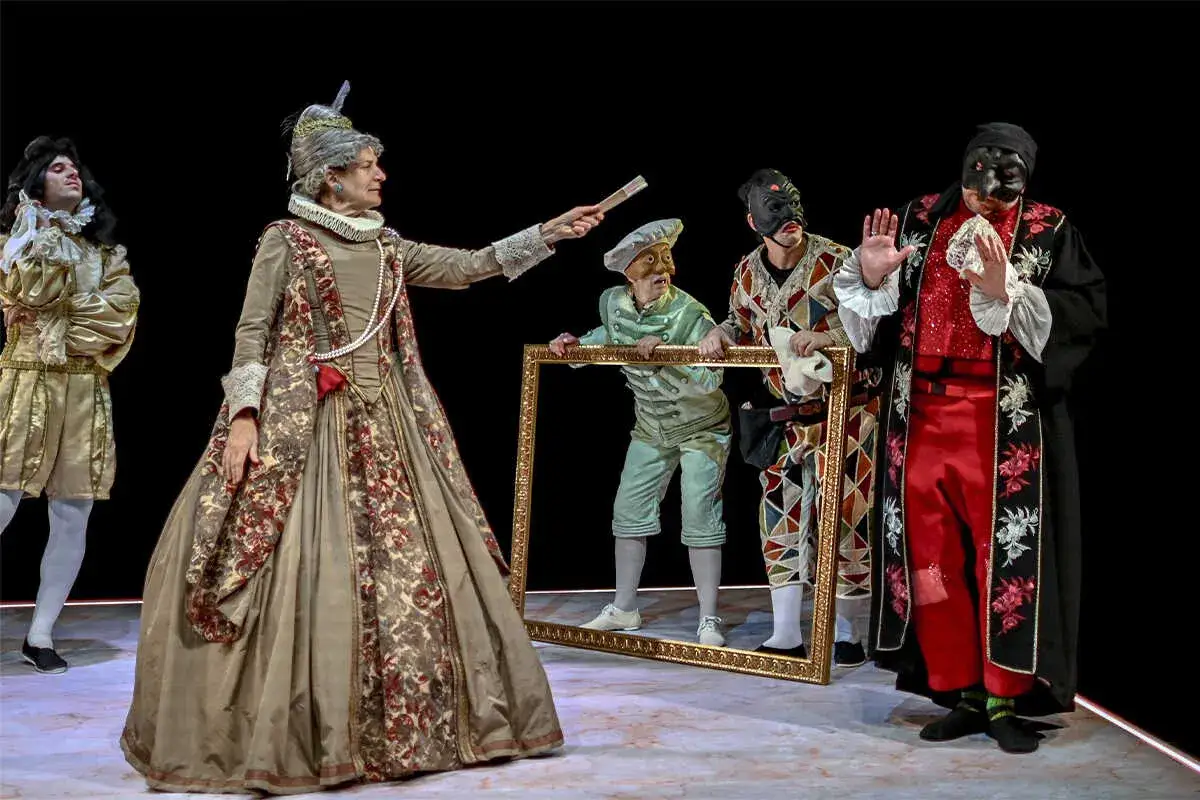 Escena teatral con personajes de **els joglars zaragoza**. Una mujer con vestido de época y abanico, un hombre con traje dorado, y otros con máscaras y ropajes coloridos.