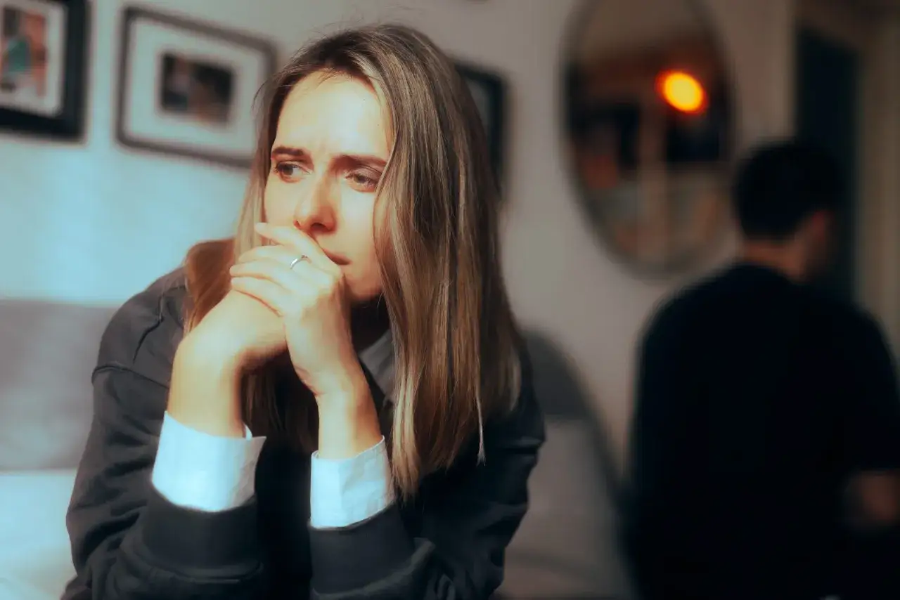 Zamyślona kobieta z rękami przy ustach, w tle niewyraźna postać mężczyzny. Czuje, że ona mnie nie kocha.