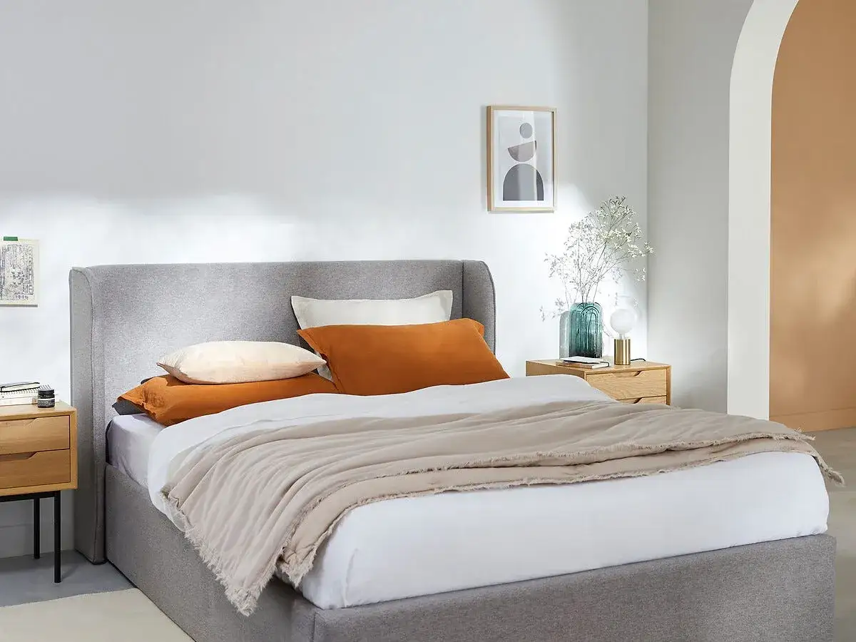 Un dormitorio moderno con un canap&eacute; gris, ropa de cama en tonos tierra y una mesita de noche de madera.