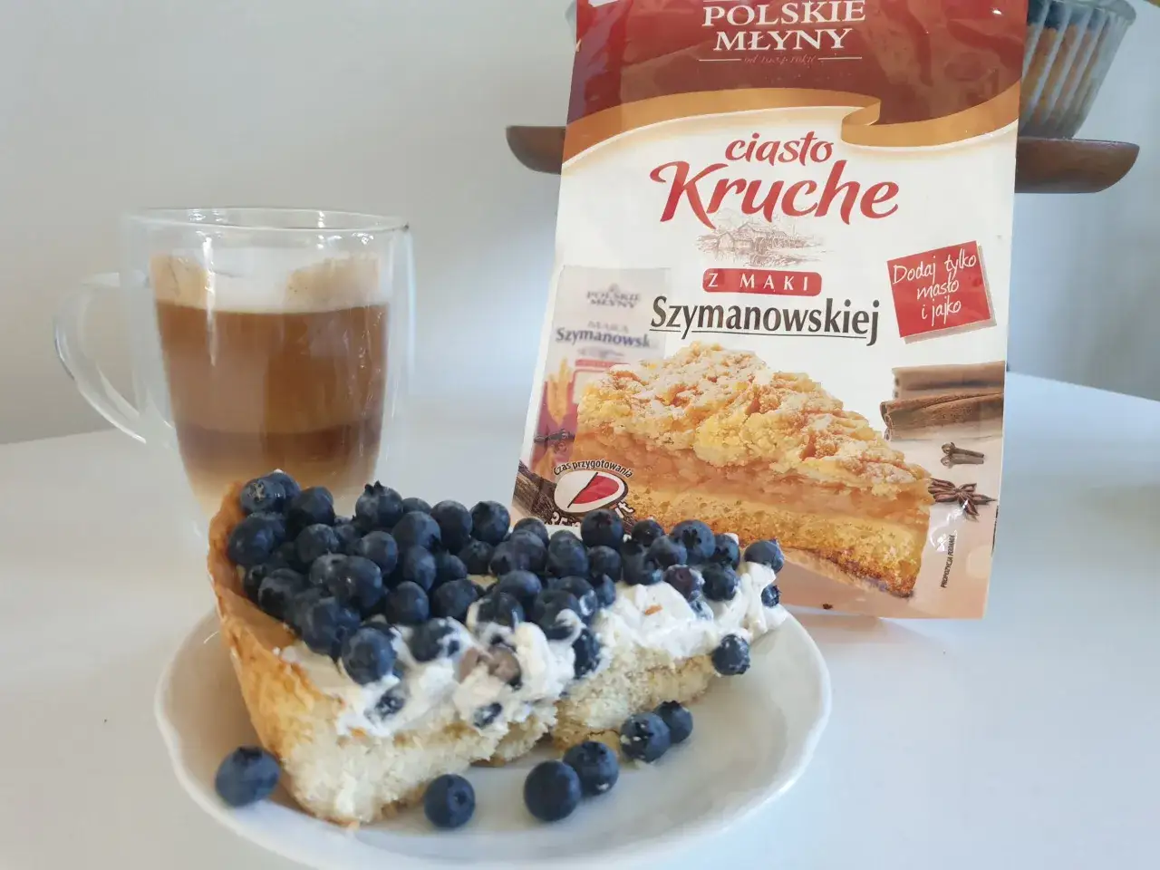 Pyszna tarta z bor&oacute;wkami i kremem, obok latte i opakowanie ciasta kruchego. Idealny deser.