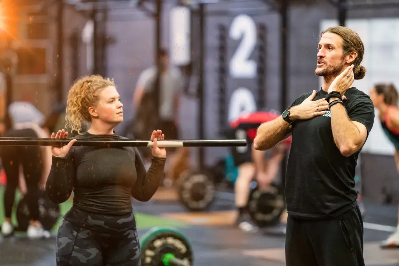 Trener wyjaśnia co to jest crossfit kobiecie z gryfem na barkach. W tle inni ćwiczący.