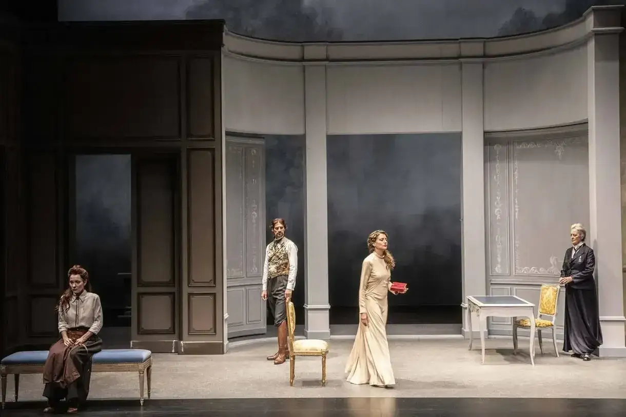 Scène de théâtre : quatre personnages dans un décor sobre. Une femme assise, un homme debout, une femme en robe longue tenant un livre, et une femme plus âgée. Une évocation des