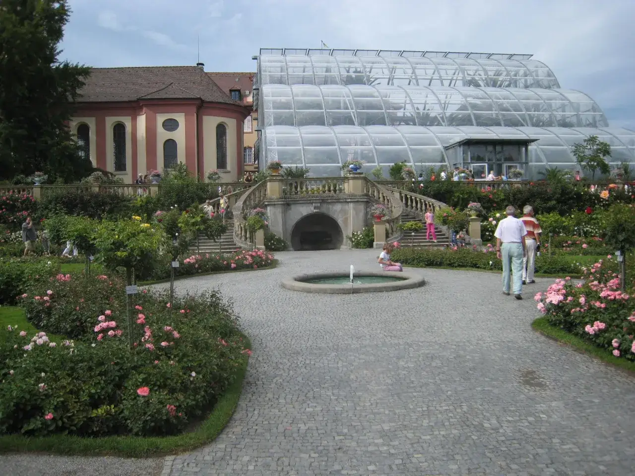 Vacker tr&auml;dg&aring;rd vid Mainau slott med rosenbuskar, en font&auml;n och en imponerande glaskonstruktion.