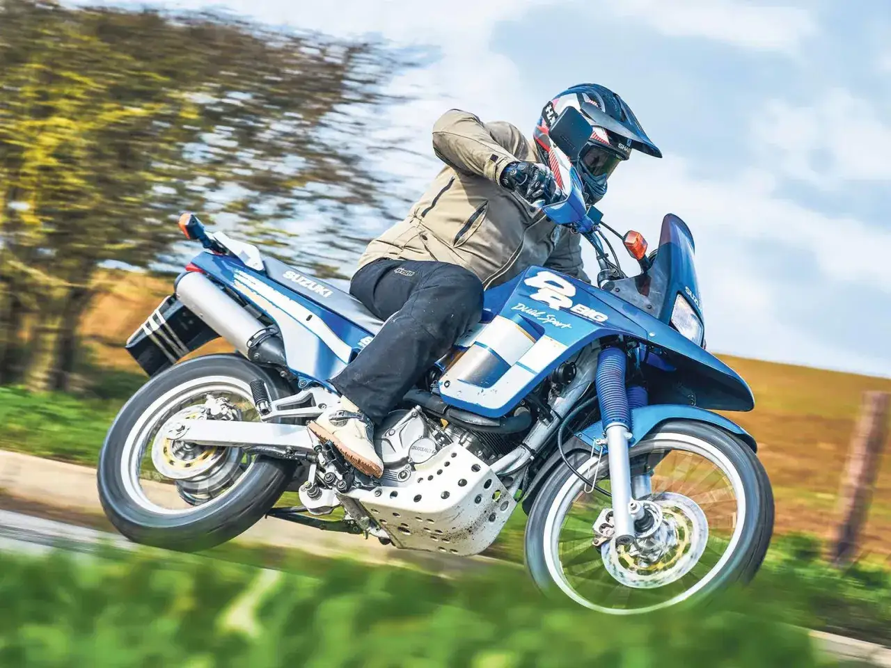 Motocyklista w kasku pędzi na niebieskim Suzuki DR 800 BIG po szutrowej drodze, otoczony zielenią i niebem.