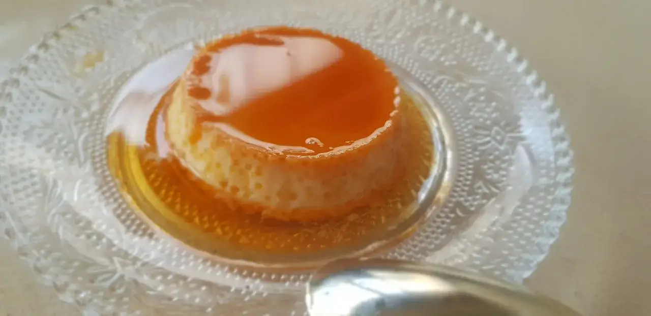 Un délicieux flan aux œufs, nappé de caramel ambré, repose sur une assiette en verre ciselé. Une cuillère argentée est prête à le déguster.