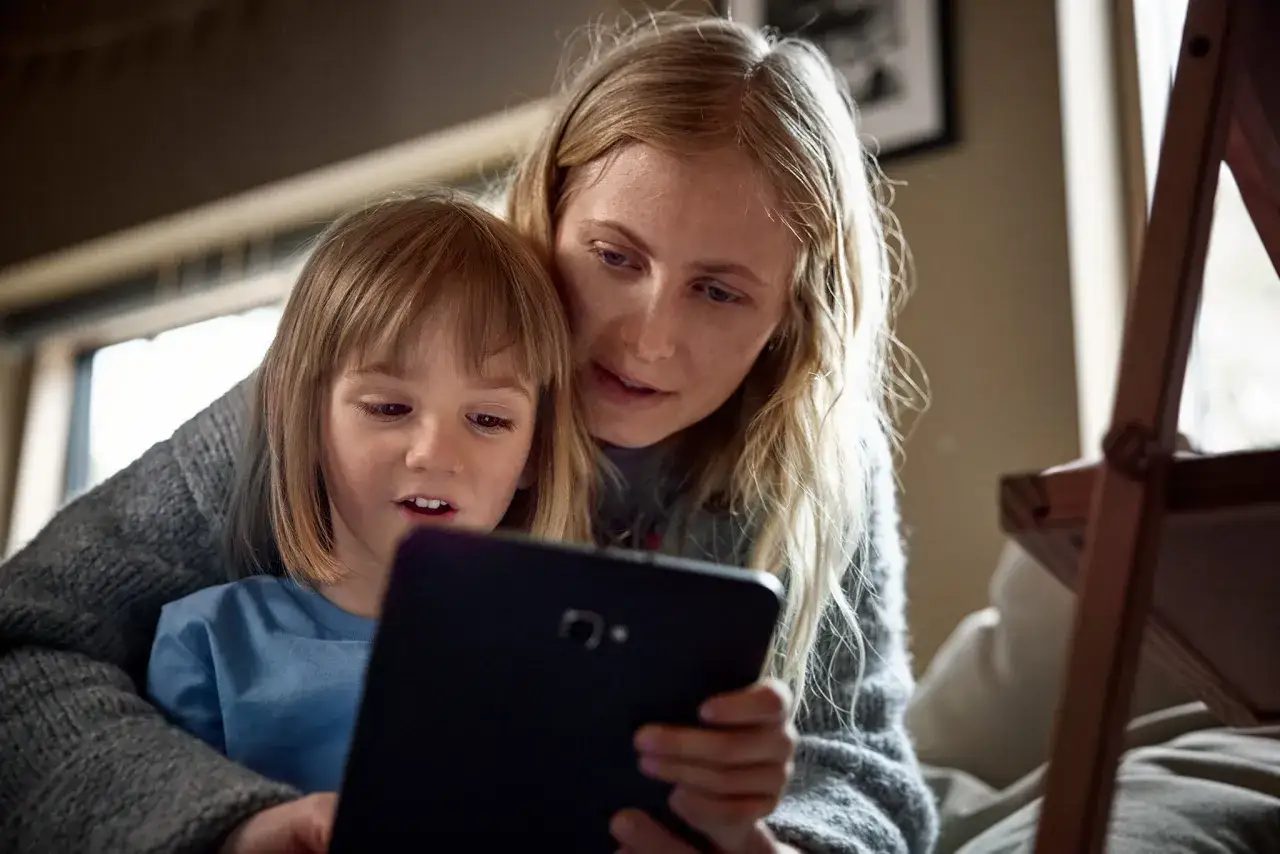 Kvinna och barn tittar på en surfplatta, kanske på sociala medier. Barnet är i förgrunden, kvinnan bakom.