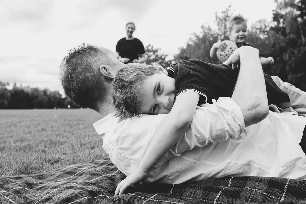 Urocze sesje zdjęciowe dzieci pomysły: chłopiec przytula się do taty na pikniku, w tle mama i brat.