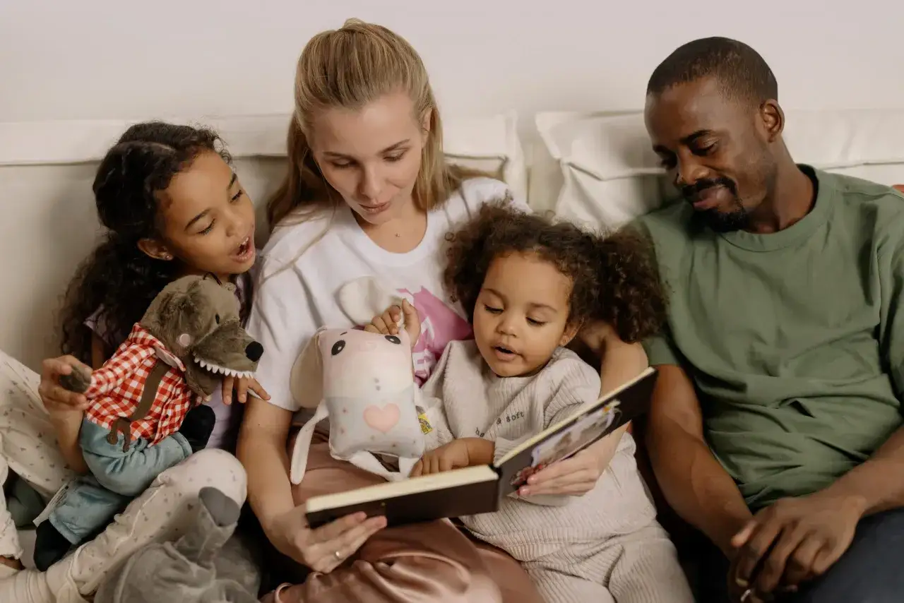 Familia reunida leyendo cuentos para ni&ntilde;os de 8 a&ntilde;os. La madre lee un libro a sus dos hijas y al padre, rodeados de peluches.