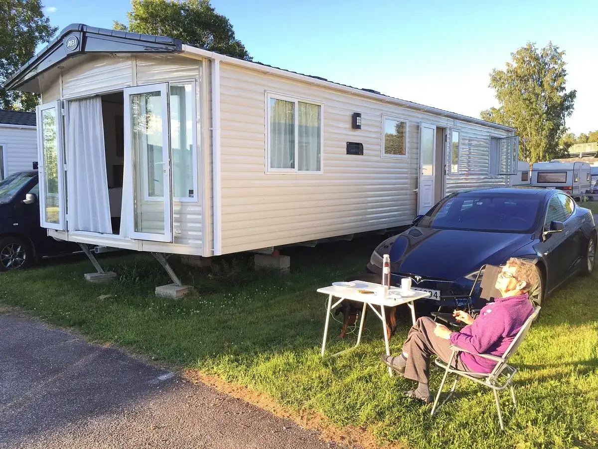 En person njuter av solen vid ett campingbord utanf&ouml;r en husvagn i Timr&aring;. En Tesla st&aring;r parkerad bredvid.