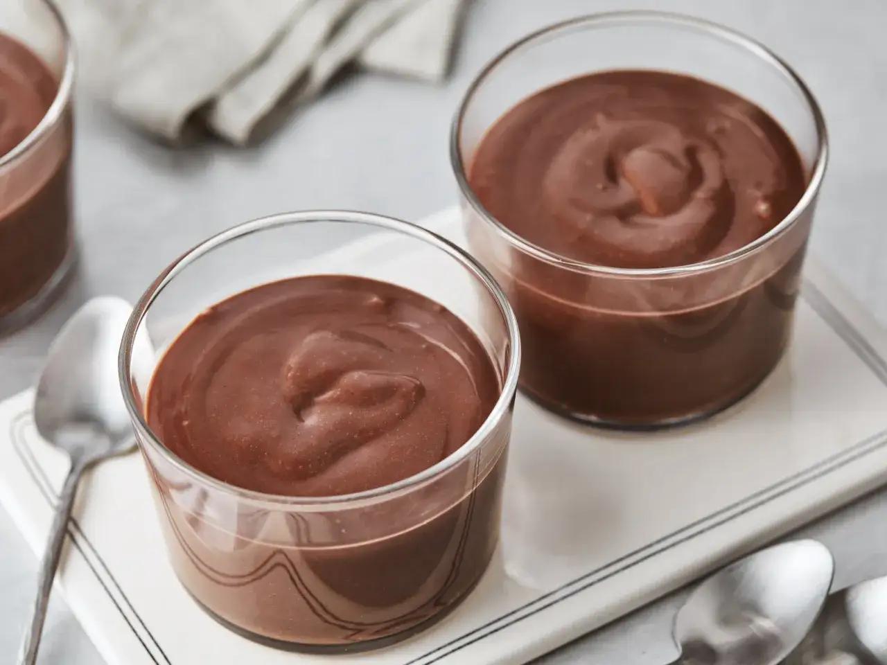 Zwei Gläser mit cremigem Schokoladenpudding, perfekt zum Pudding selber machen. Löffel liegen bereit.