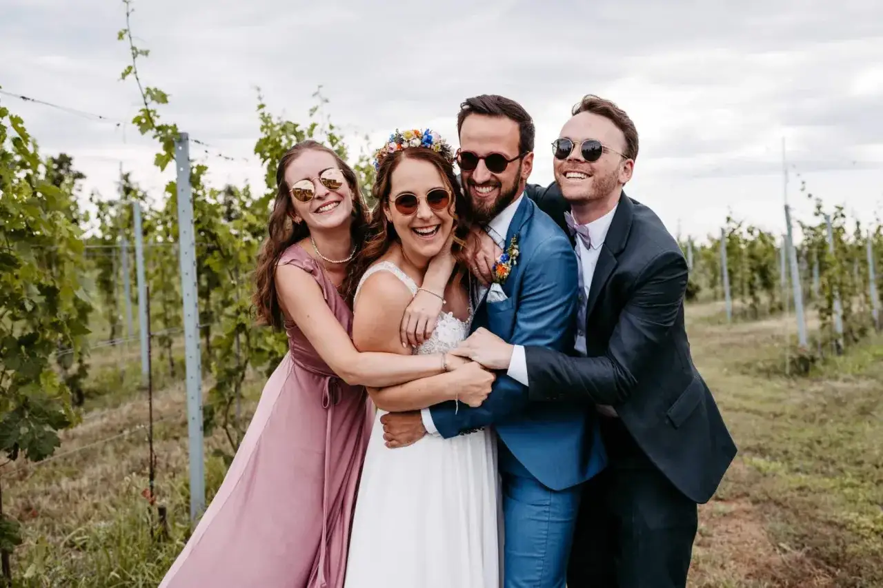 Gl&uuml;ckliches Brautpaar mit seinen **Hochzeit Trauzeugen** in einem Weinberg, alle tragen Sonnenbrillen und umarmen sich fr&ouml;hlich.