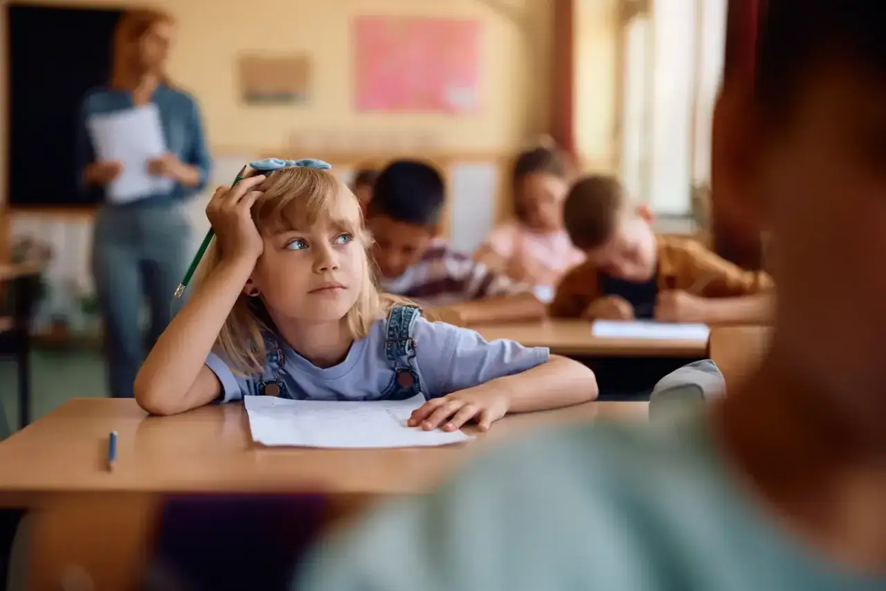 Dziewczynka z blond włosami i niebieskimi oczami, oparta o rękę, z oł&oacute;wkiem w dłoni, zastanawia się nad lekcją. Czy jej średnia na 3?