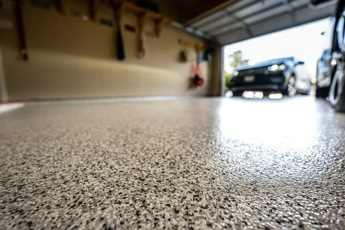 Un sol de garage fraîchement peint, avec des flocons gris et noirs, reflétant la lumière. Des voitures sont garées à l'extérieur, la porte du garage est ouverte.
