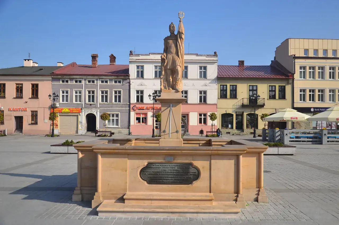 Pomnik na rynku w Żywcu. Kamienna fontanna z rzeźbą żołnierza na cokole, otoczona kolorowymi kamienicami.