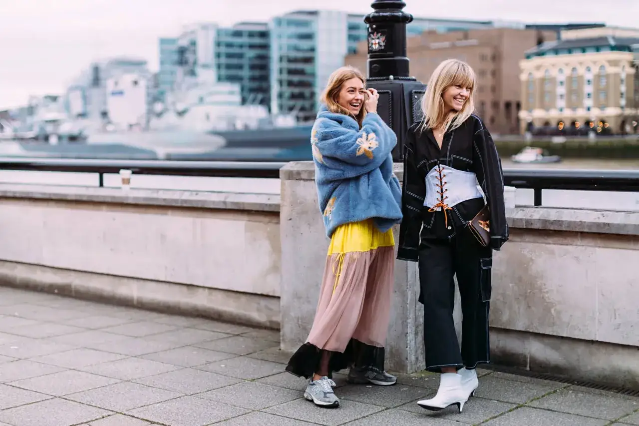 Dwie kobiety w modnych stylizacjach, jedna w niebieskim futerku i zwiewnej sukience, druga w czarnym kombinezonie z gorsetem. Obie noszą buty New Balance, co stanowi świetny new balance poradnik.