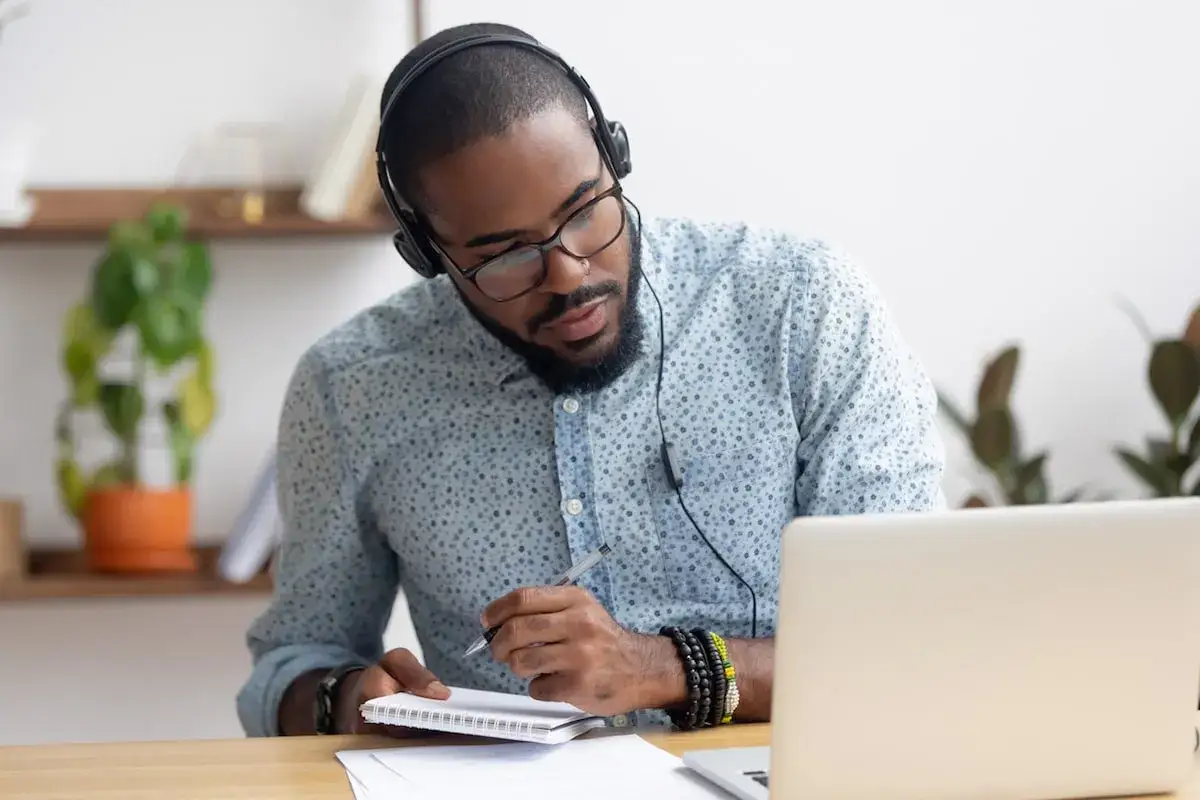Hombre con auriculares y gafas, concentrado, escribiendo en un cuaderno. Est&aacute; practicando franc&eacute;s con su port&aacute;til.
