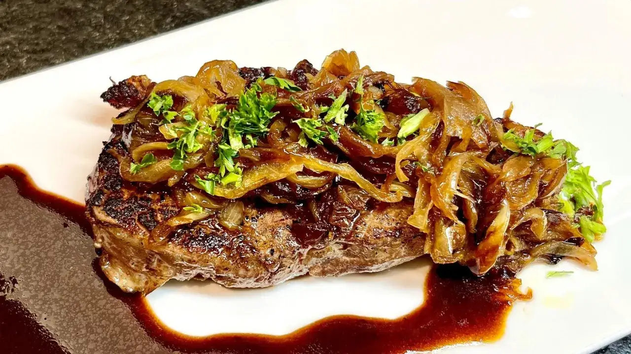 Ein saftiger Rostbraten, welches Fleisch wohl? Mit karamellisierten Zwiebeln und frischer Petersilie garniert, serviert auf einer weißen Platte mit dunkler Sauce.