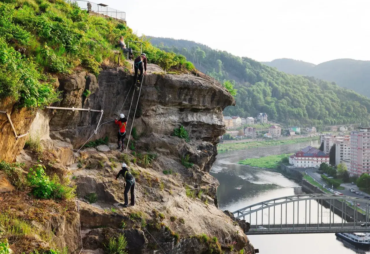 Osoby wspinają się po skalnej ścianie na via ferrata w Děčínie. Widok na rzekę i miasto.
