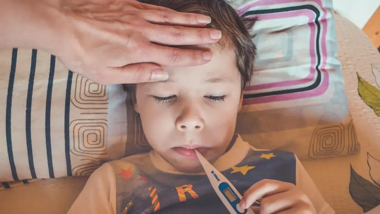 Niño con termómetro en la boca y mano en la frente, indicando dolor de cabeza y fiebre en niños.