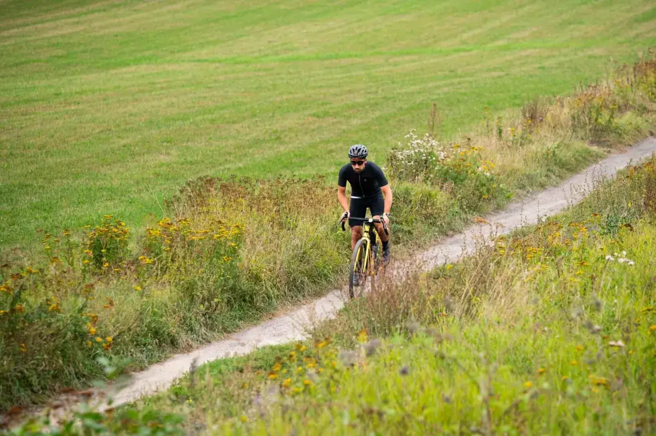 Mężczyzna na rowerze przemierza polną drogę wśród zielonych łąk i polnych kwiatów. To idealne miejsce na Mazury rowerem.