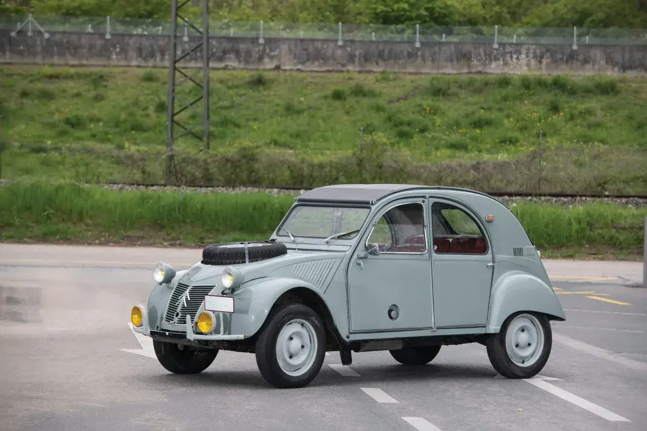 En ljusgr&aring; Citro&euml;n 2CV st&aring;r parkerad vid en v&auml;g med gr&ouml;nskande natur i bakgrunden.