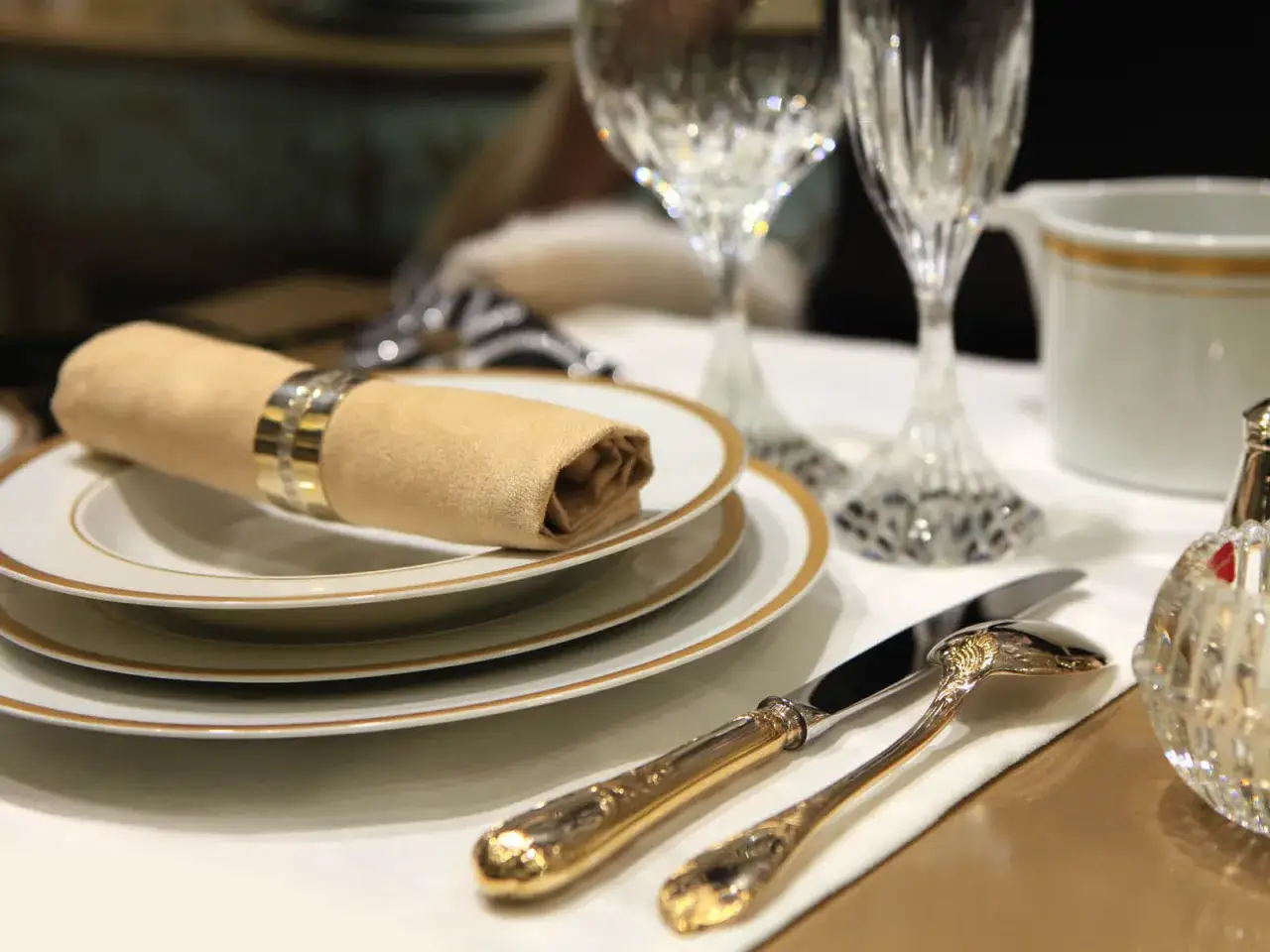 Une table élégamment dressée, évoquant le raffinement du dressage à la française. Serviette beige, couverts dorés, verres ciselés.