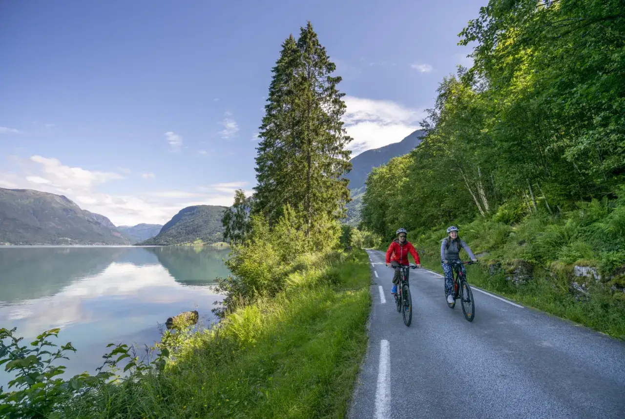 Två personer cyklar längs en väg vid en spegelblank sjö i Norge. Berg och grönskande skog omger dem.