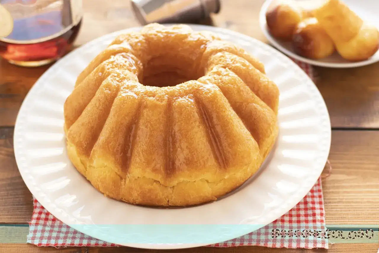 Un soffice bab&agrave;, pronto per essere inzuppato, evoca la **bab&agrave; ricetta originale**. Accanto, un bicchierino di rum e altri bab&agrave; in miniatura.