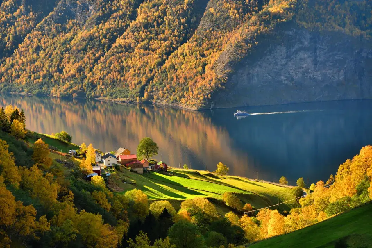 Jesienny fiord w Norwegii, z kolorowymi drzewami na zboczach gór i małą wioską nad wodą. Statek płynie po spokojnej tafli.