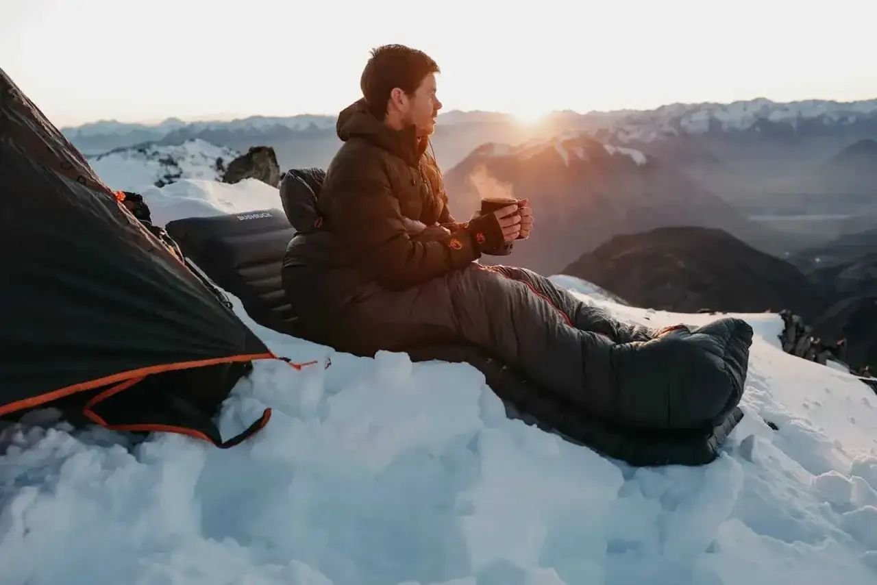 Mann genießt heißen Tee im Schlafsack auf verschneiter Bergspitze. Die 600 cuin Füllung hält ihn warm, während die Sonne über den Gipfeln aufgeht.