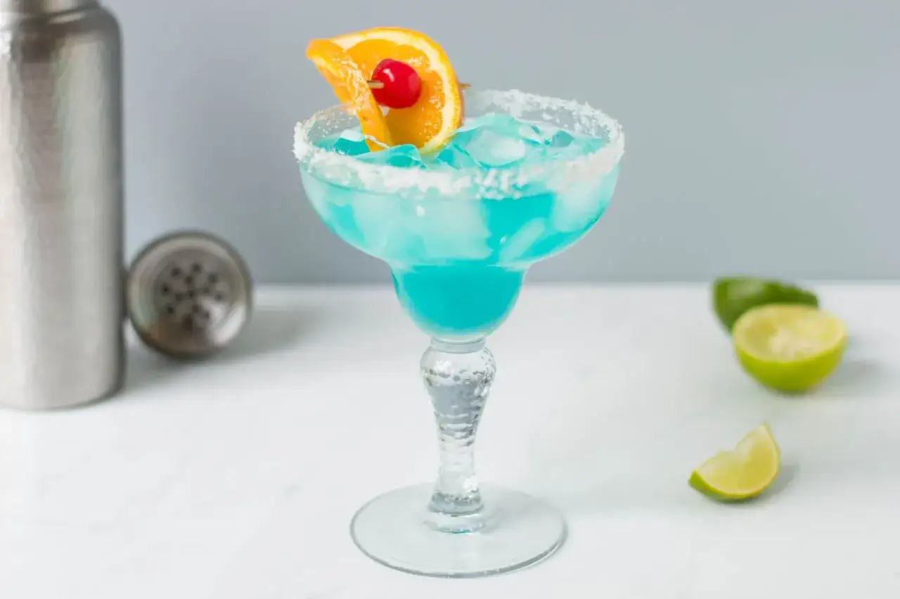 Niebieski drink nazwa, orzeźwiający koktajl w kieliszku z solą, ozdobiony plasterkiem pomarańczy i wiśnią. Obok leżą połówki limonki.