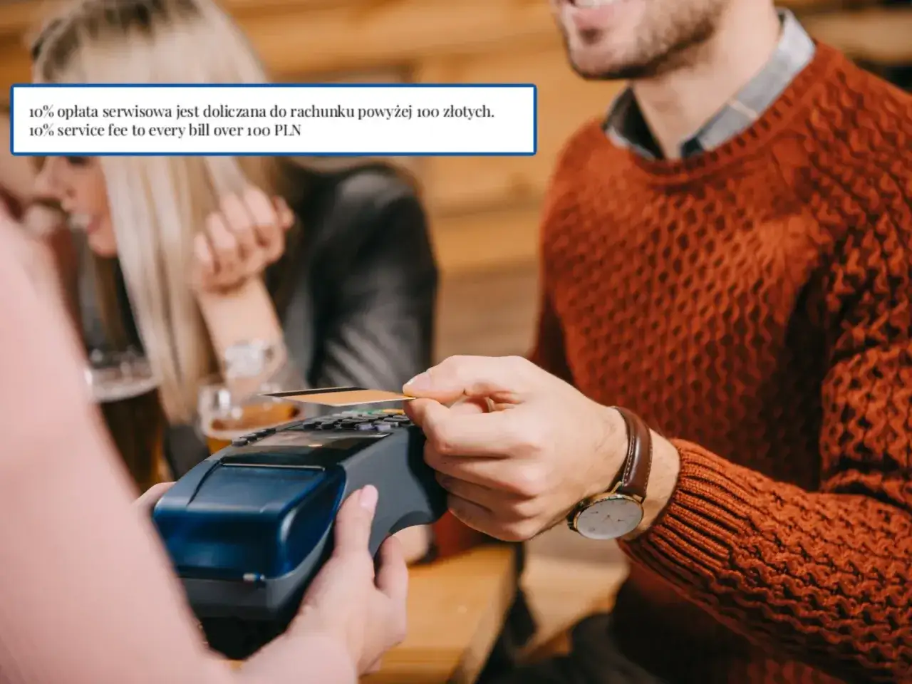 Płacenie kartą w restauracji. Na ekranie terminala widać informację o 10% opłacie serwisowej do rachunku powyżej 100 zł.