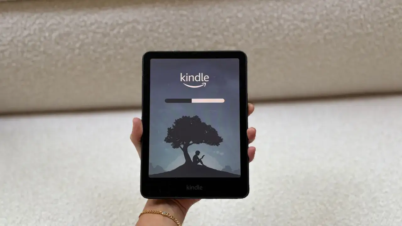 Une main tient une liseuse Kindle affichant un &eacute;cran de chargement avec un arbre et une silhouette lisant. L'utilisateur s'appr&ecirc;te &agrave; &eacute;teindre le Kindle.
