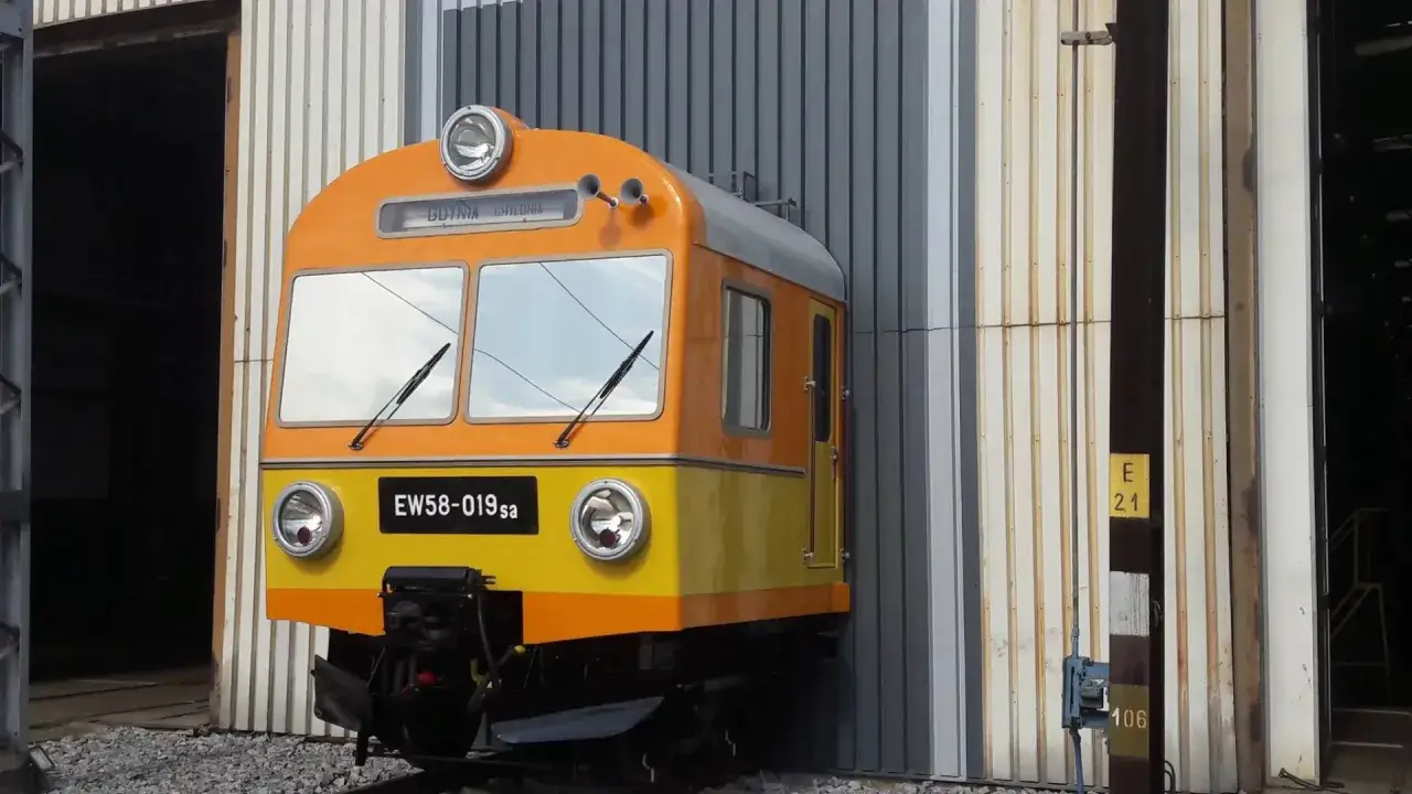 Pomarańczowo-żółty pociąg EW58-019sa stoi przed halą.
