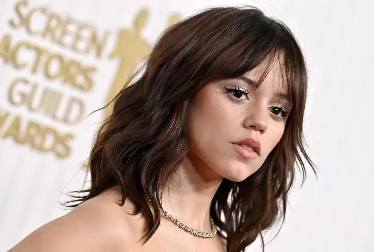 Jenna Ortega z brązowymi włosami, które tworzą stylowe **face framing**, w białej sukni i z błyszczącym naszyjnikiem.