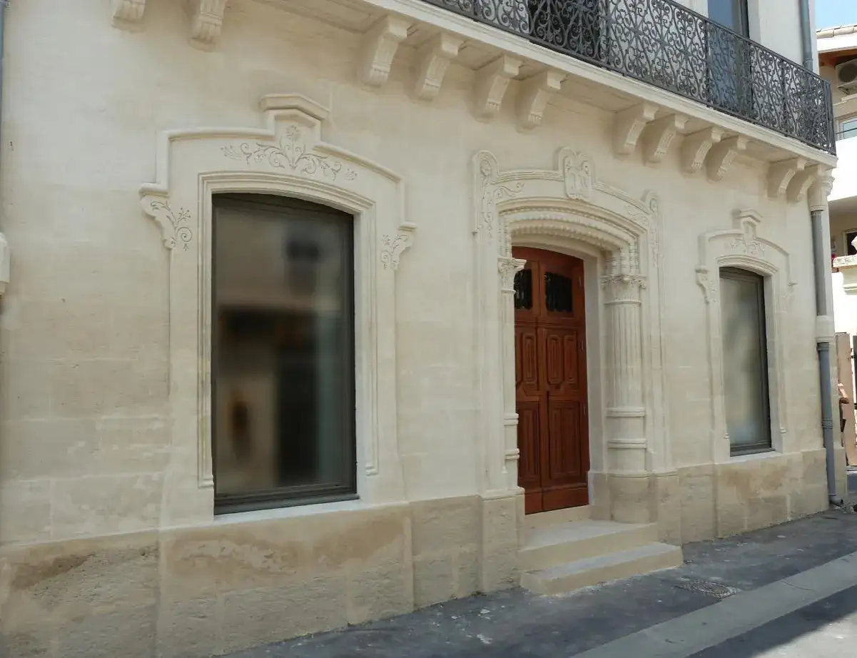Façade en pierre de taille, avec une porte en bois sculptée et des fenêtres ornées. La chaux, utilisée pour la finition, met en valeur les détails architecturaux.