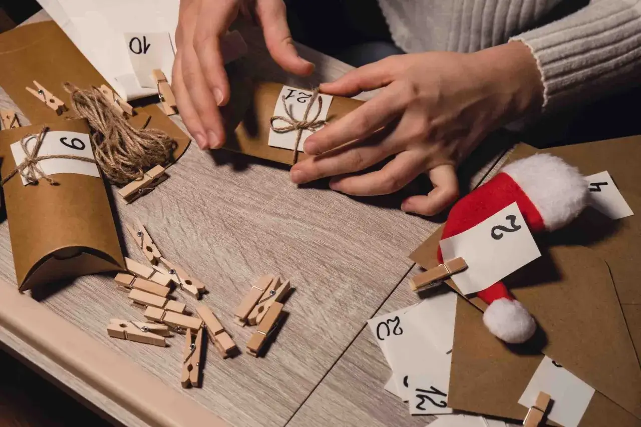 H&auml;nder som pysslar med ett julkalender diy. Sm&aring; bruna paket med siffror och kl&auml;dnypor ligger utspridda.