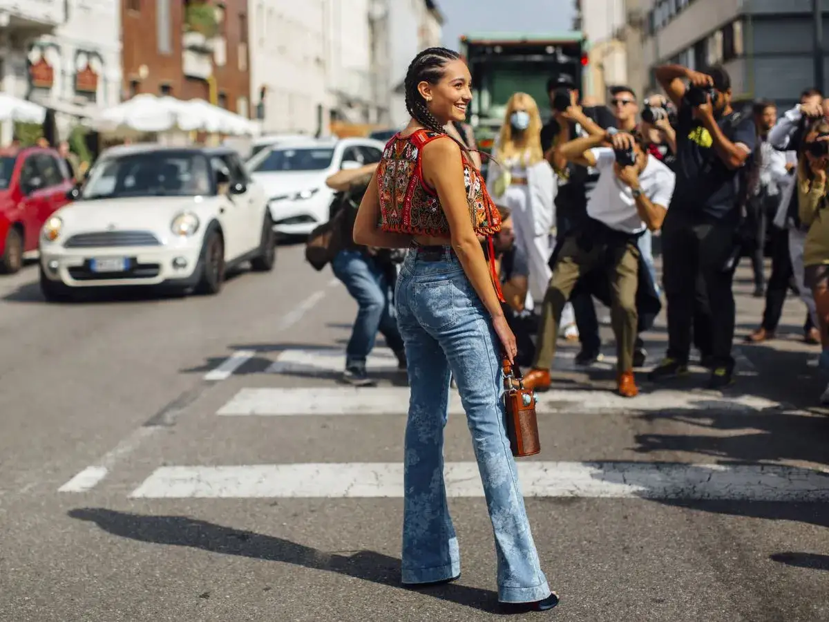 Mujer con trenzas y top bordado, pantalones acampanados y bolso de mano, luciendo un auténtico estilo bohemio mientras es fotografiada en la calle.