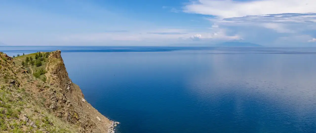 Skalisty brzeg z zielenią i ścieżką prowadzi do bezkresnego, błękitnego jeziora. W oddali majaczą góry. To malowniczy port nad Angarą.