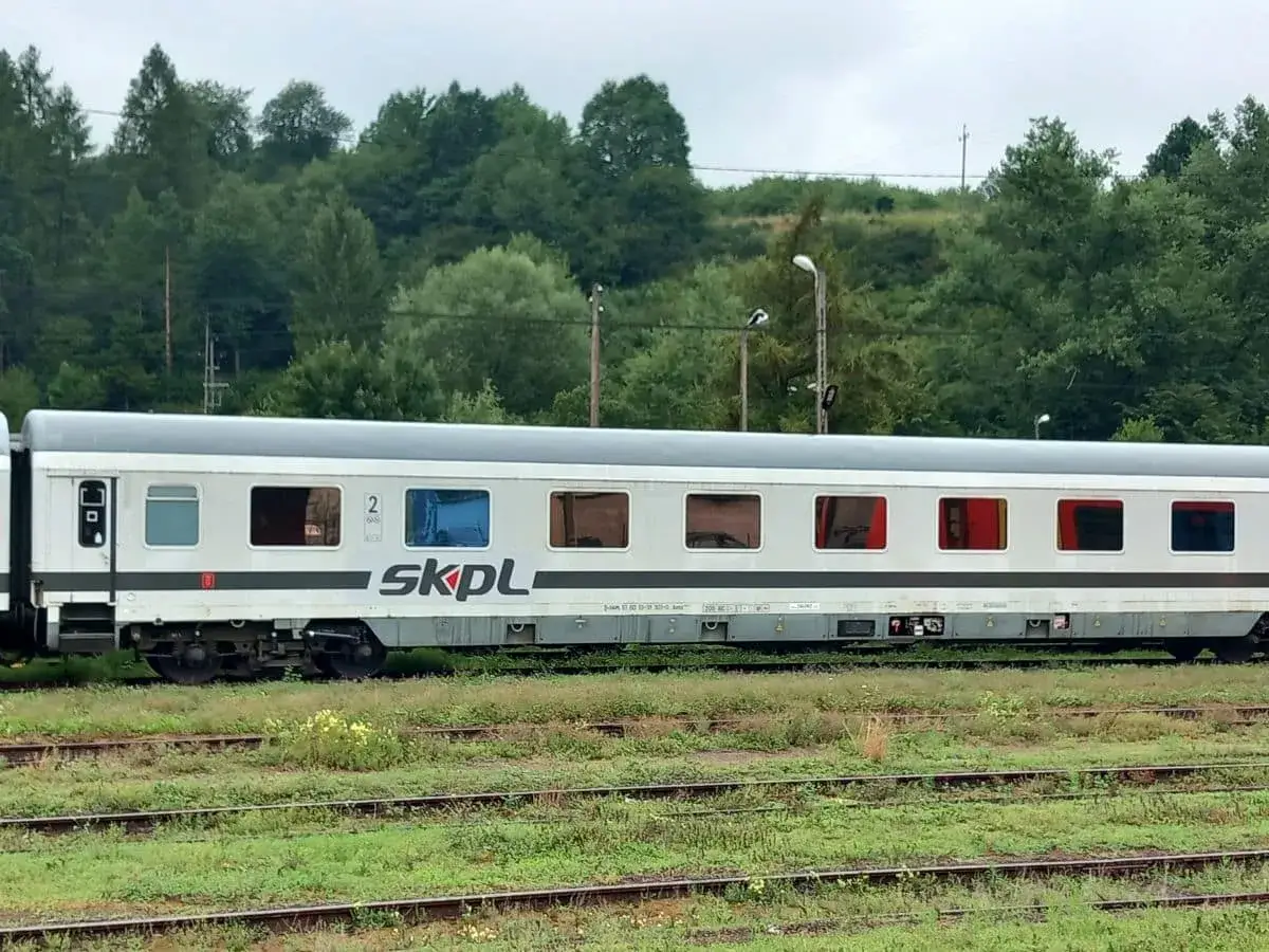 Biały wagon PKP z napisem SKPL, stojący na torach wśr&oacute;d zieleni.