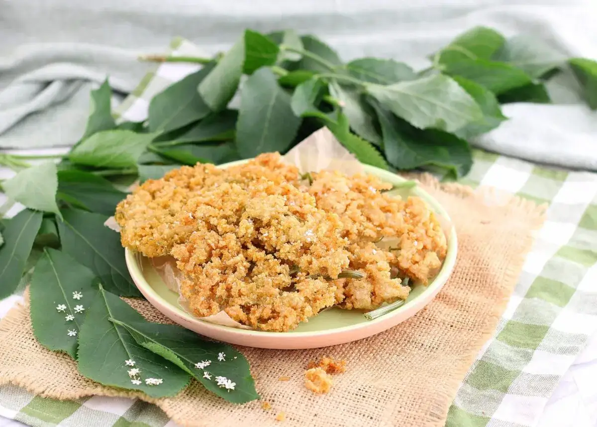 Deliziosi fiori di sambuco fritti, dorati e croccanti, serviti su un piatto con foglie verdi e piccoli fiori bianchi.