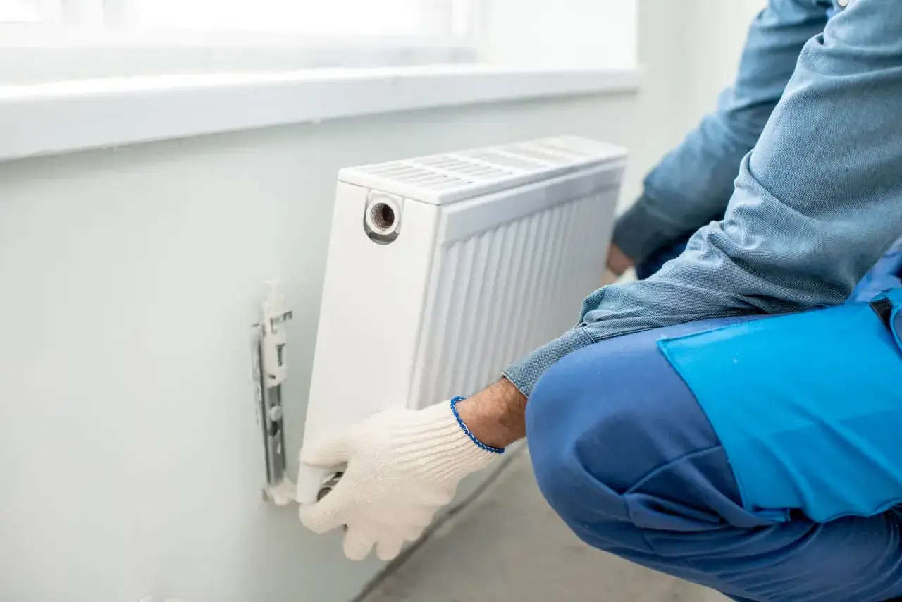 Man con guantes blancos ajusta un radiador blanco en la pared. Se está realizando la tarea de cambiar radiador de sitio.