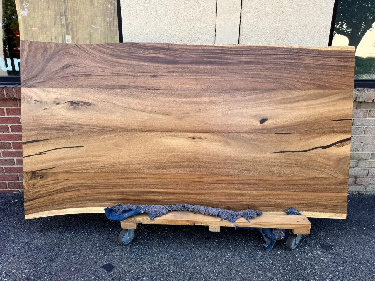 A large, live-edge acacia lumber slab with beautiful, swirling grain patterns rests on a wooden dolly.